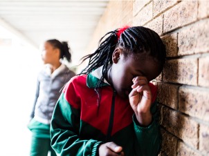 A school girl stands leaning against a wall crying, she is being bullied by other school girls.